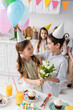 © LIGHTFIELD STUDIOS - happy girl in party cap holding tulips near boy and next to friends during birthday party at home.