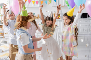 group of joyful kids in party caps dancing under falling confetti during birthday celebration at home.