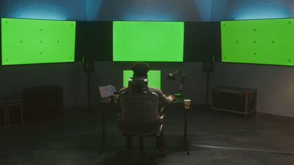 Fototapete - African American man works on computer in modern studio with equipment. Multiple green screen big monitors on the wall. Professional software displayed on tablet. Color grading or film editing room.