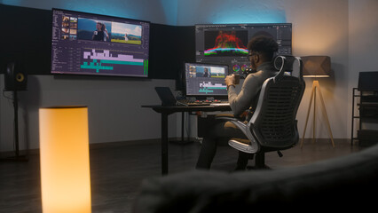 Fototapete - African American multimedia artist drinks coffee and makes color grading in editing program on computer. Process of video post production in modern studio. Multiple monitors with travel movie footage.