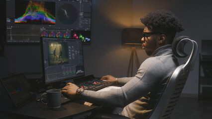 Fototapete - African American film maker makes color grading on computer in modern studio. Big screen with RGB colour correction graphic bar on wall. Action film footage displayed on monitor. Colorist equipment.