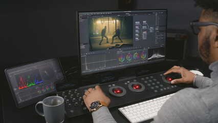 Fototapete - African American colorist makes color grading on control machine in modern studio. Big screens with action film footage and RGB colour correction graphic bar on the wall. Film post production process.