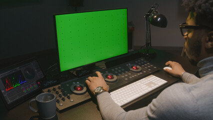 Fototapete - African American film maker works on computer in studio with digital equipment. Color grading control surface and tablet with professional software standing on the table. PC monitor with green screen.
