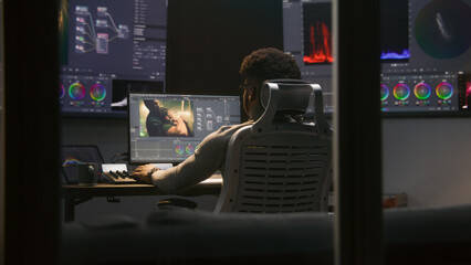 Fototapete - Experienced African American video maker does color grading in program on computer. Process of colour correction for movie post production in modern studio. Multiple monitors with action film footage.