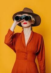 Poster - Stylish blond hair woman in hat and brown dress with binoculars on yellow background