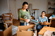 © Drazen - Happy Asian woman and her female roommate unpacking cardboard boxes while moving into new apartment.
