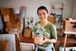 © Drazen - Happy Asian woman holds potted plant while moving into new home and looking at camera.