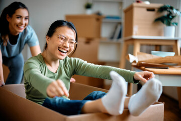 Wall Mural - Young Asian woman laughs while her friend is pushing her in cardboard box at their new home.