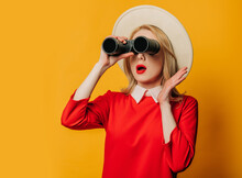 Surprised Blonde Hair Woman In Hat And Red Dress With Binoculars On Yellow Background