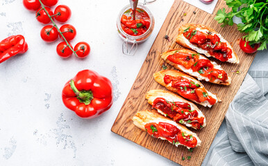 Bruschetta with soft cheese and grilled red paprika in olive oil with herbs on cutting board, white kitchen table background, top view