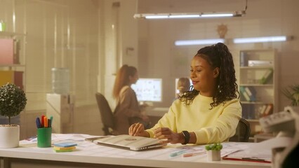 Young african american woman is closing her laptop and texting on her mobile phone. Black female programmer or designer in yellow sweatshirt orders food, communicates on Internet, browses information.