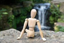 A Wooden Artist's Figure Sitting On A Rock Looking At A Waterfall. Concept Of Reflecting In Nature
