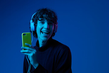 Wall Mural - Man smile with phone in hand taking selfies with headphones listening to music, portrait dark blue background, neon light, style and trends, mixed light, men's fashion, copy spot