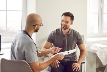 Portrait of a happy smiling young man patient in medical office listening to doctor holding report file with appointment and giving consultation during medical examination in clinic.