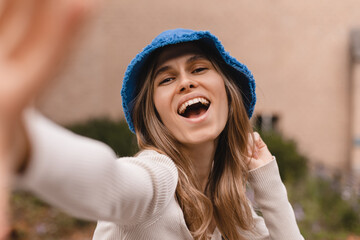 Wall Mural - Close up young caucasian woman look at camera, hold her panama hat, spends her leisure time on walk in street. Brunette wears blue panama hat, top with long sleeves. Girl make selfie photo, open mouth