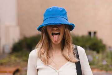 Wall Mural - Attractive blonde woman wear bright blue panama hat and hide her eyes by cap, show tongue and laughing. Joyful woman look happy and funny, funky. Girl in sweater top and bag on shoulder.