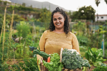 happy indian woman smiling on camera in the garden while holding basket of fresh vegetables - harves