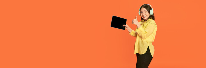 portrait of smiling, young, positive, asian girl in headphones, holding tablet against orange studio