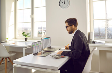 male accountant working on two modern laptops. young man sitting at office desk, doing paperwork, lo