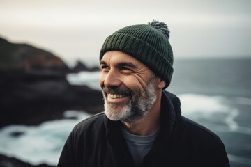 Wall Mural - Portrait of a smiling bearded man in a hat and jacket standing on the seashore at sunset