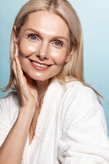 Wall Mural - Vertical shot of smiling mid 50s woman with aging wrinkles, touches her clear glowing skin on face, applies skincare toners and cosmetic lotion, wears bathrobe on blue background