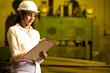 © GAJENDRRA BHATI  - Indian female engineer plant supervisor wearing safety hard hat holding paper and pad working in industrial factory. skill india and women empowerment concept.