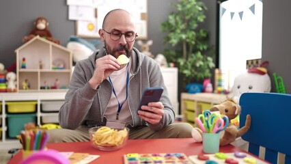 Wall Mural - Young bald man preschool teacher using smartphone eating chips potatoes at kindergarten