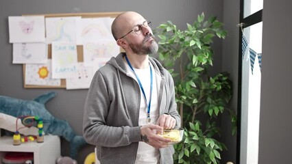 Canvas Print - Young bald man working as teacher eating chips potatoes at kindergarten