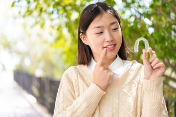 Wall Mural - Young Chinese woman at outdoors holding invisible braces with happy expression