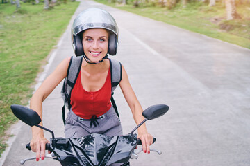 Wall Mural - Tropical travel and transport. Young smiling woman in helmet riding scooter on the road