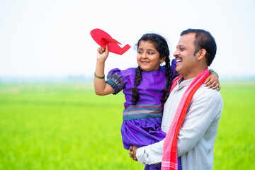 Wall Mural - Happy village girl kid playing with airplane toy while father holding or carrying near farmland - concept of relationship, parental caring and healthy lifestyle