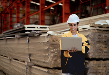 Female industrial engineer in white helmet and safety jacket work in heavy metal engineering factory. Latin technician woman worker using laptop computer in metalwork manufacturing facility, portrait.