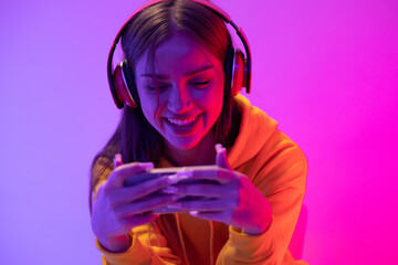 Shot of a young adult female wearing headphones, plying games, listening to music. Keeping in touch with others in a joyful and confident gesture. Modern studio with vibrant neon lighting.