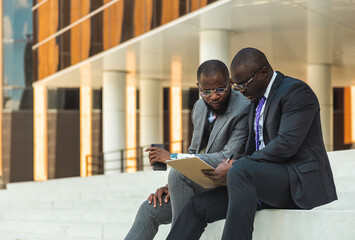 Sticker - Friendly meeting of business partners outdoors. Two dark-skinned men in suits sit on the steps of a city building with a notebook and have a conversation. Working break. Support and collaboration