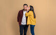 Side view of girlfriend hiding her mouth and telling secret to surprised boyfriend. Young couple sharing gossips with each other while standing isolated on beige background