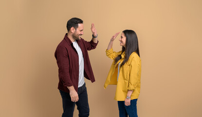Wall Mural - Smiling young couple in shirts giving high-five to each other and celebrating victory. Cheerful man and woman clapping hands and greeting while standing on isolated beige background