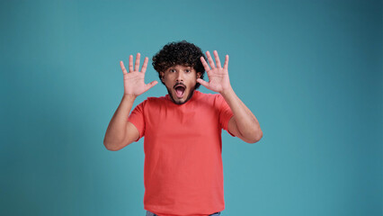 Horror, thriller, scare story. Portrait with young frightened indian man , guy screaming and raising hands with fear face in coral t-shirt on blue studio background. Human emotions, psychological 