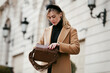 © Anna Zhuk - Beautiful young woman opening purse bag and taking something out while standing on street against of building. Stylish female model with fashionable hairstyle