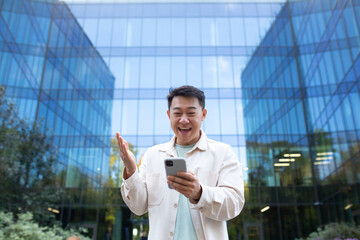 Portrait of a young Asian man standing near a glass skyscraper and holding a phone. Happy received good news, reads messages, chats on video call.