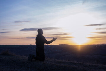 A man with raised hands is praying. On the background of the sunset sky. Repentance for sins. Kneeling Prayer to God. Worship and praise.