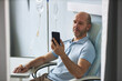 © Seventyfour - Portrait of bald adult man using smartphone during IV drip treatment in clinic, copy space