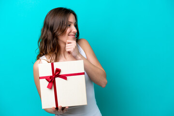 Wall Mural - Young caucasian woman holding a gift isolated on blue background thinking an idea and looking side