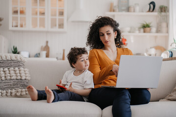 Smart young Italian woman with curly hair dressed in orange blouse and blue pants sitting on sofa using laptop, working at home at the kitchen with son playing on phone, talking with mom. Family