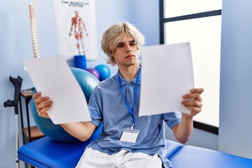 Poster - Young blond man pysiotherapist reading document sitting on massage board at rehab clinic