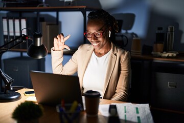 Sticker - Beautiful black woman working at the office at night smiling and confident gesturing with hand doing small size sign with fingers looking and the camera. measure concept.