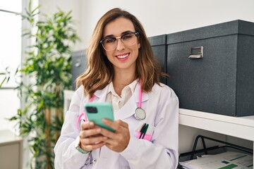 Canvas Print - Young woman wearing doctor uniform using smartphone at clinic