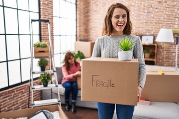 Canvas Print - Mother and daughter moving to a new home holding cardboard box winking looking at the camera with sexy expression, cheerful and happy face.