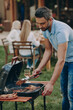 © gstockstudio - Confident young man barbecuing meat on grill while his family relaxing in the background