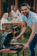 © gstockstudio - Happy young man barbecuing meat on grill while his family relaxing in the background