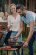 © gstockstudio - Happy young couple barbecuing meat on grill while their family relaxing in the background